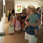Woman holds small child while they book look at paintings on the wall. Other children can be seen in the background learning about art from a staff member.