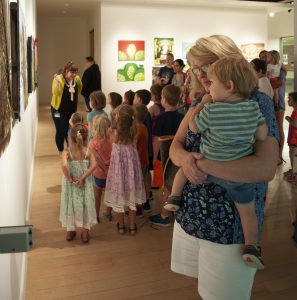Woman holds small child while they book look at paintings on the wall. Other children can be seen in the background learning about art from a staff member. 