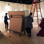 Museum staff roll works of art into the galleries on an A-frame dolly. We only see the backs of several canvases, not the actual artwork.