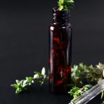 A small, long glass brown bottle sits with some herbs coming out of it and a dropper pipette next to it.
