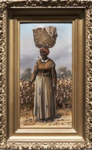 988.001.0207_edited Female African American figure in a worn dress and a red kerchief standing in a cotton field, balancing a large basket of cotton on her head. She is smoking a corncob pipe and holds a walking stick in her proper right hand.