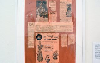 Framed newspaper clippings of John Meigs and an old advertisement for Sears featuring illustrations of women and girls in Aloha themed dresses and tops