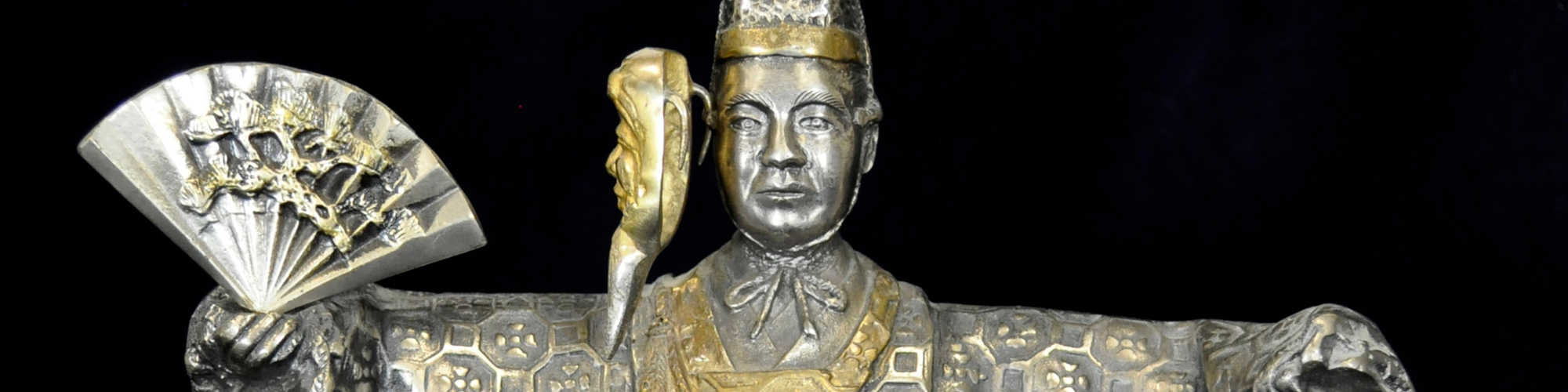 Color photograph of a brass, male Japanese theatre figure in an elaborate layered winter kimono with gold mask and fan. Mask is hinged to 'open' and reveal the actor's face.