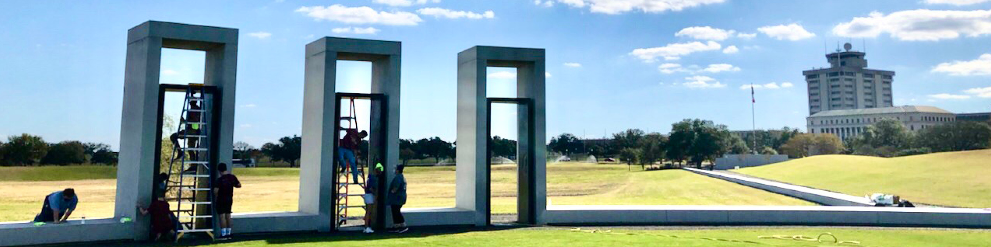 Color photograph of students on ladders cleaning the portals at the Bonfire Memorial Site. The Administration and Oceanography/Meteorology Buildings are in the right background. 
