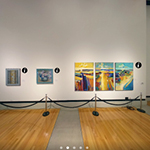 360 degree view of the interior of an art gallery. Colorful paintings hang on the wall at a distance and there is a sculpture on a pedestal on the far left. 
