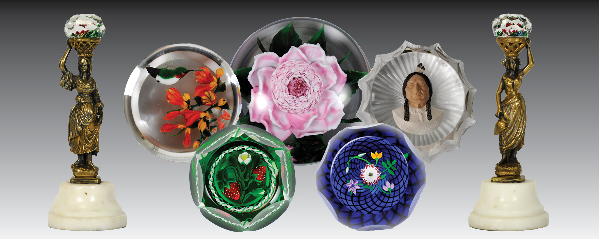 Various paperweights with glass objects inside: a hummingbird, a pink cabbage rose, Sitting Bull, strawberries, and flowers. Two statues of women holding baskets on their heads with a glass orb inside flank the paperweights. 