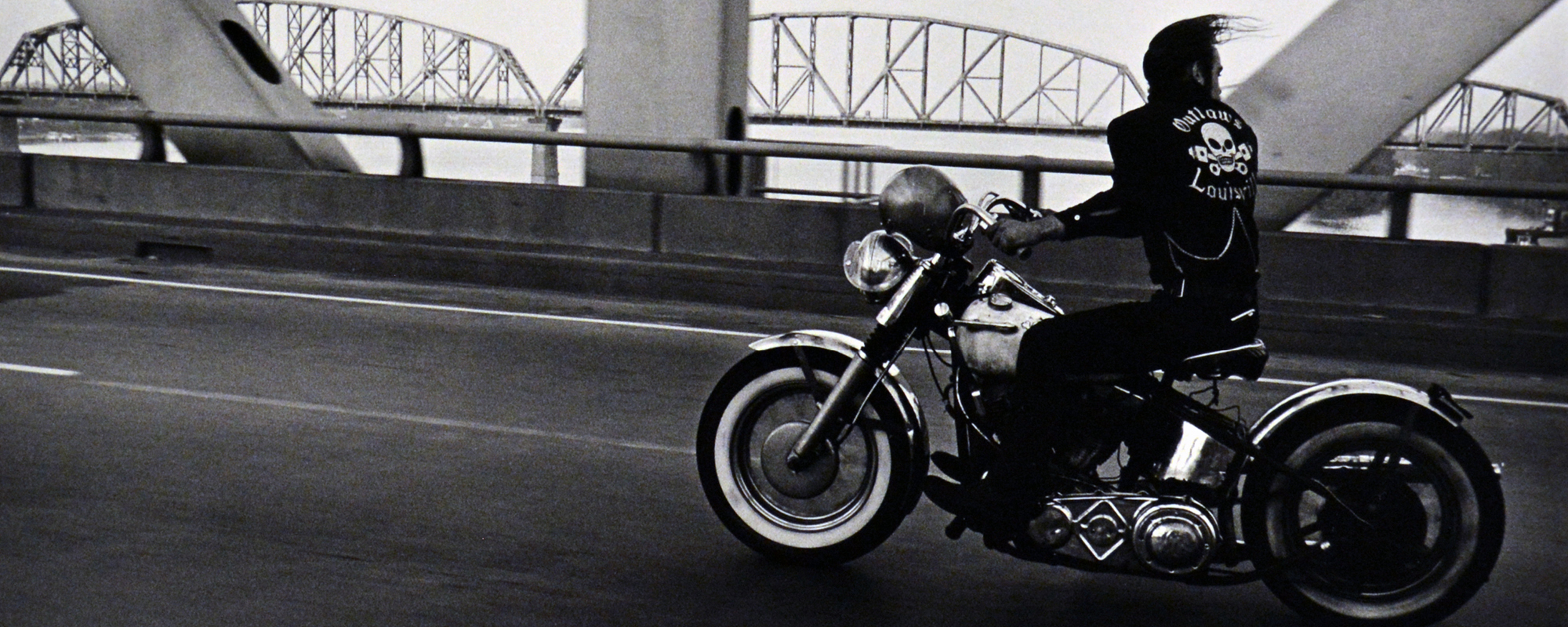 Person in leather jacket riding motorcycle across bridge over water. 