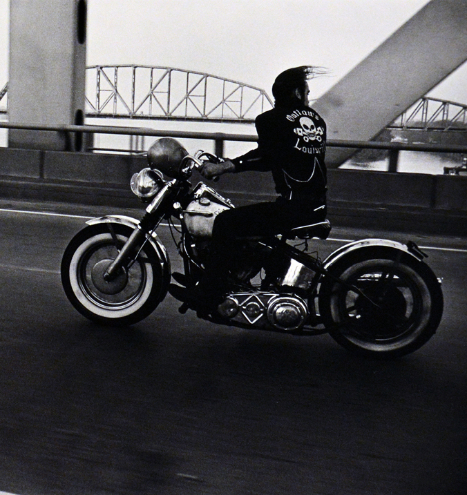 Person in leather jacket riding motorcycle across bridge over water.