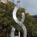 Menos tall, silver tone sculpture standing tall outdoors with trees behind it.