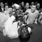 Black and white photograph of Big Jay McNeely laying on stage playing saxophone on his back. An audience of teenagers looks on in exuberance.