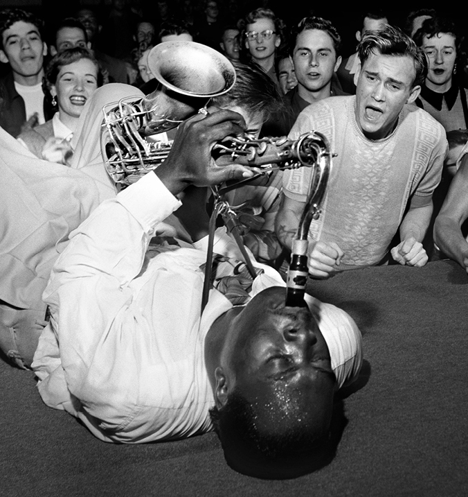 Black and white photograph of Big Jay McNeely laying on stage playing saxophone on his back. An audience of teenagers looks on in exuberance. 