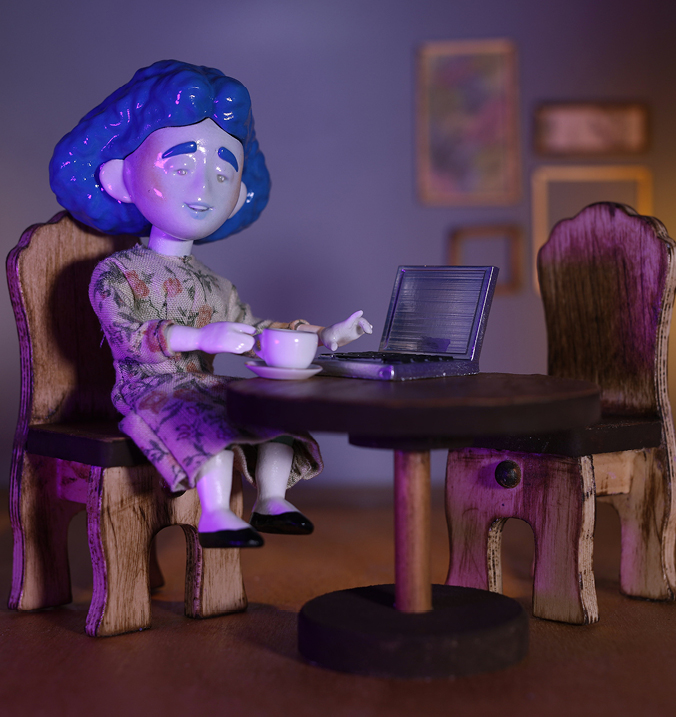 Tiny porcelain doll figurine sits at tiny wooden table and chairs with a saucer cup in one hand and the other on a tiny laptop.