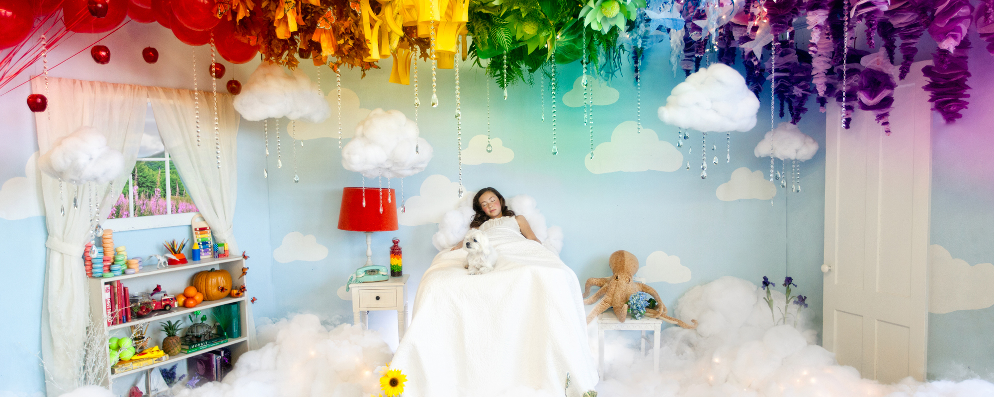 A young girl sleeps in a white bed with rainbow colored paper clouds above her.