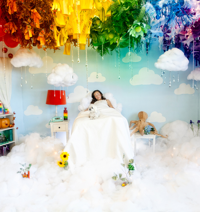 A young girl sleeps in a white bed with rainbow colored paper clouds above her.