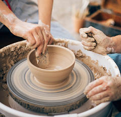 Two sets of hands work on a clay bowl on a potter wheel.